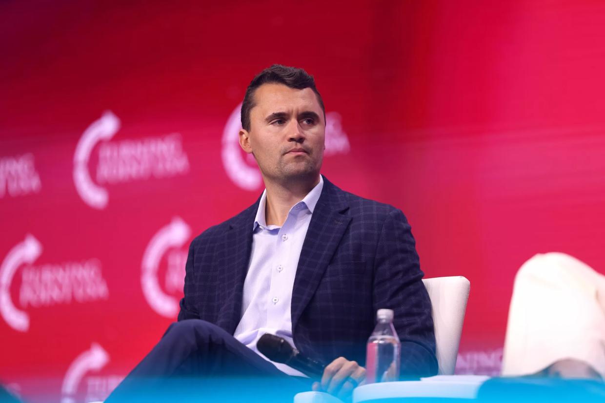color photo of a man wearing a white open collared shirt, navy blue sport coat, and navy blue pants seated at a public event against a magenta background